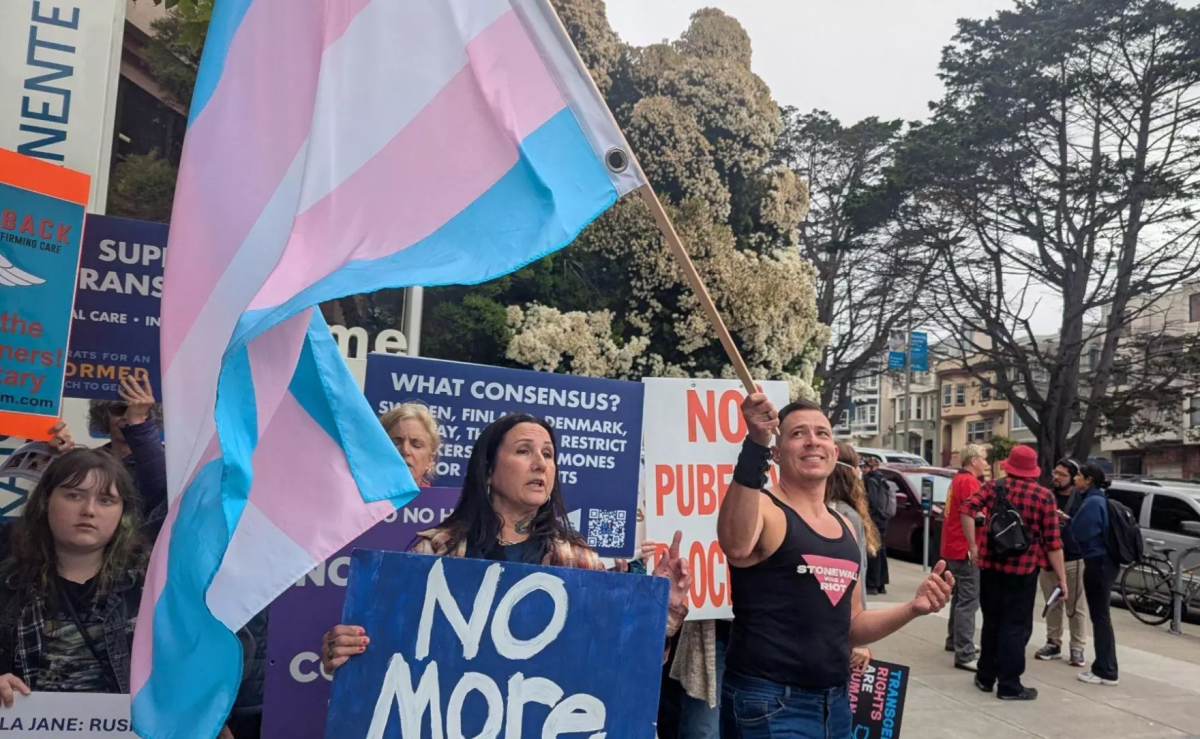 Calder Storm waves a trans flag over a crowd of counterprotestors at a rally for trans youth outside of a Kaiser Permanente clinic in San Francisco on July 25, 2025.